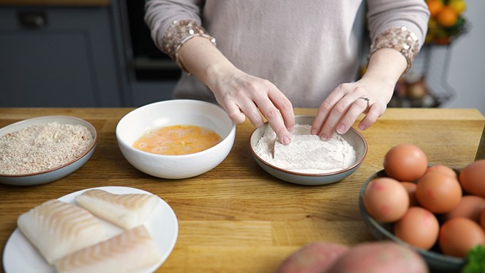 Hands dipping a haddock fillet into a bowl of flour. Bowl of whisked egg and a further bowl with breadcrumbs in scene. Plate of 3 haddock fillets and bowl of eggs partially in shot.