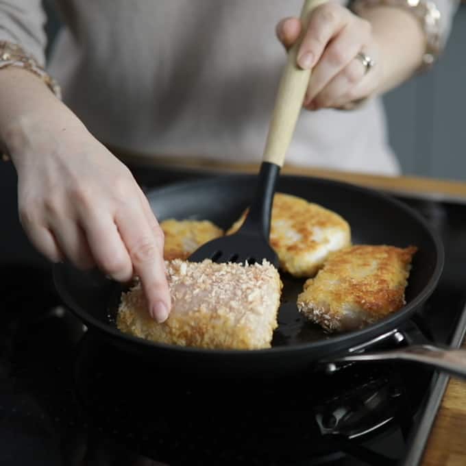 Frying pan with 4 breaded haddock fillets frying. One is being turned over with a spatula.