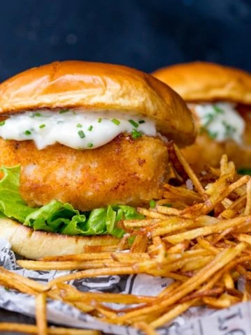 Breaded haddock burger on brioche bun with lettuce and chive-speckled mayonnaise. Shoestring fries to the right of the burger. A second burger in background.