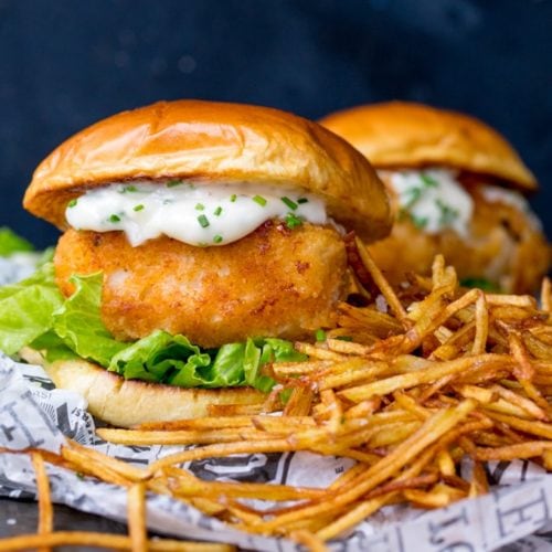 Breaded haddock burger on brioche bun with lettuce and chive-speckled mayonnaise. Shoestring fries to the right of the burger. A second burger in background.