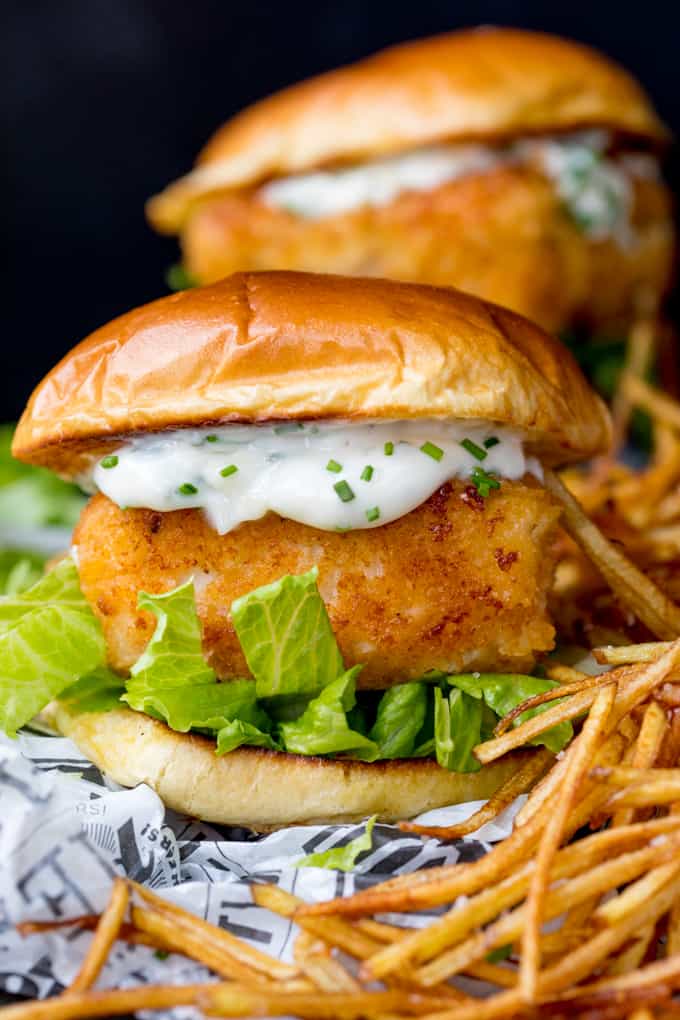 Close up image of breaded haddock burger on brioche bun with lettuce and chive-speckled mayonnaise. Shoestring fries to the right of the burger. A second burger in background.