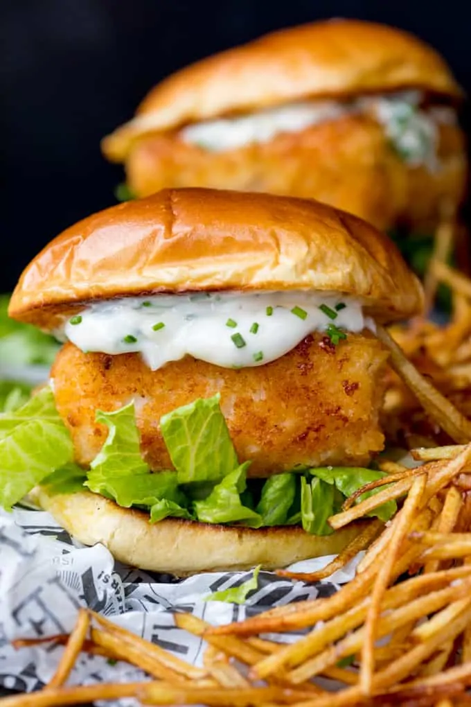 Close up image of breaded haddock burger on brioche bun with lettuce and chive-speckled mayonnaise. Shoestring fries to the right of the burger. A second burger in background.