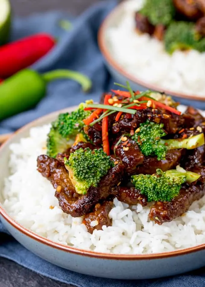 Crispy beef strips and broccoli in a tangy sauce on top of a bowl of boiled rice. Further bowl in the background.
