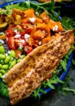 Close up image of salad bowl filled with roasted veg, tomatoes, edamame, red onion, courgette and topped with honey mustard trout.