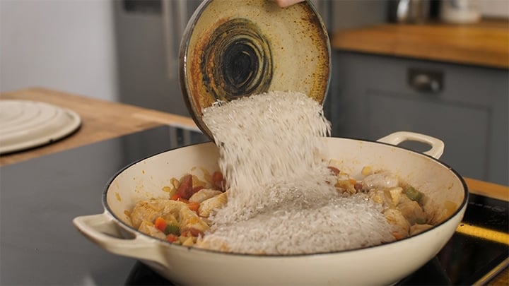 Rice being poured into a pan of chicken and chorizo for jambalaya