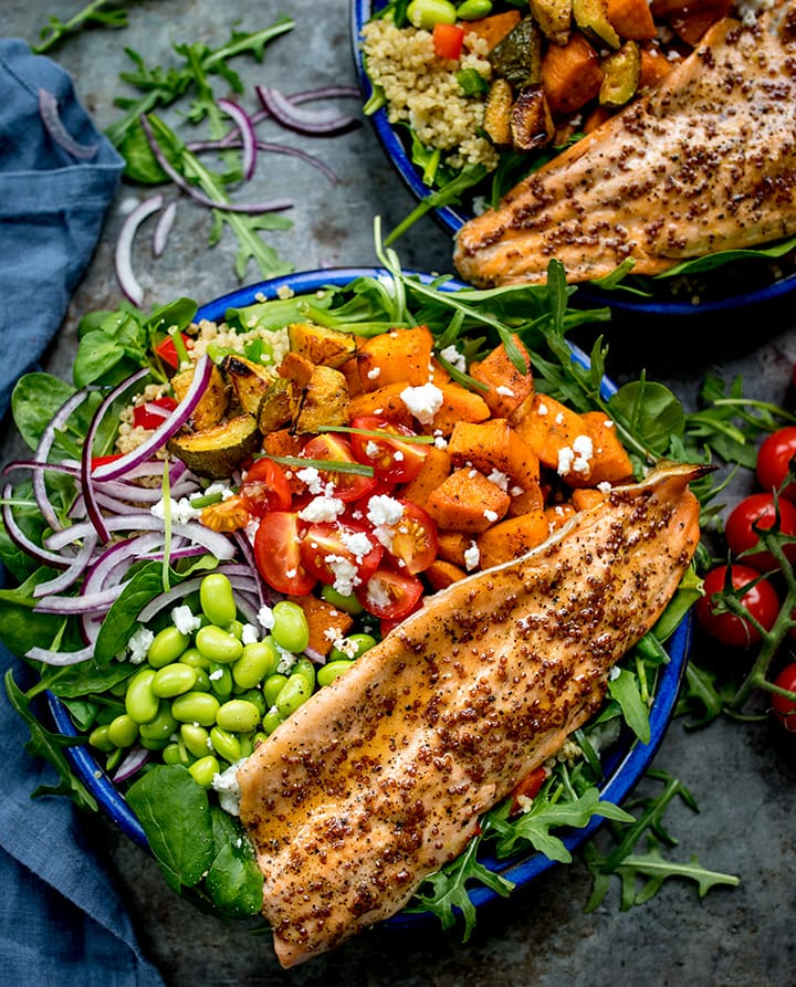 Blue bowl filled with roasted veg, tomatoes, edamame, red onion, courgette and topped with honey mustard trout.