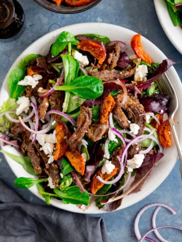 A white plate filled with crispy lamb salad with tomato, feta and red onion. The plate is on a blue background and there is another plate, salad dressing and sliced of red onion scattered around in shot.