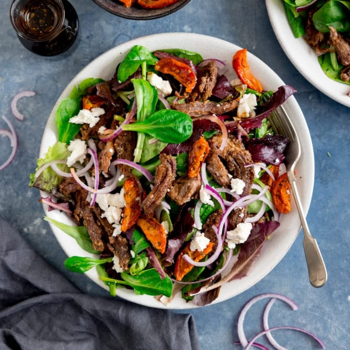 A white plate filled with crispy lamb salad with tomato, feta and red onion. The plate is on a blue background and there is another plate, salad dressing and sliced of red onion scattered around in shot.