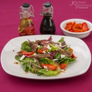 white plate with lamb, tomato and feta salad. Oil and vinegar in background next to a small bowl of sun blush tomatoes