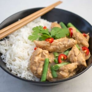 Black bowl filled with rice and peanut butter chicken curry with green beans. Chillies and coriander on top.