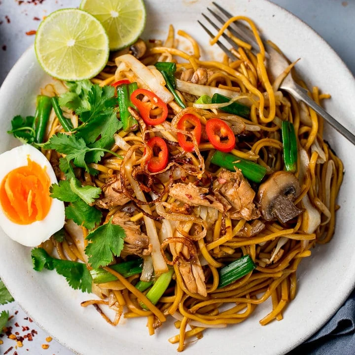 overhead of a Mee goreng (asian noodles) on a white plate with egg, chillies and lime slices