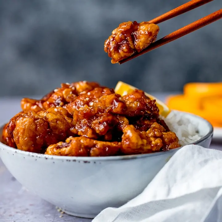 Square image of Sticky Orange Chicken Stir Fry in a bowl with boiled rice. Sliced oranges in the background.