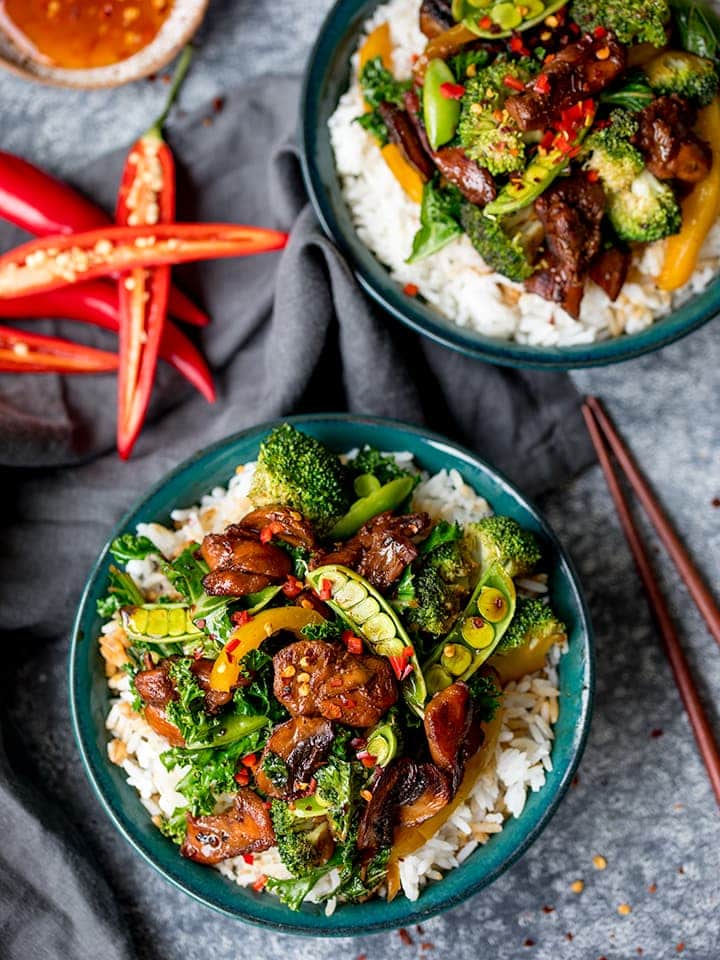 Chicken and vegetable stir fry on rice in green bowl. Sliced chillies and sweet chilli sauce next to bowl