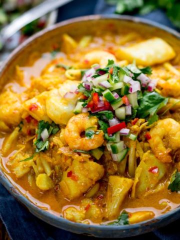 Square image of haddock and prawn laksa curry in a bowl with topping of chillies, cucumber and coriander. On a blue napkin, spoon on left hand side.