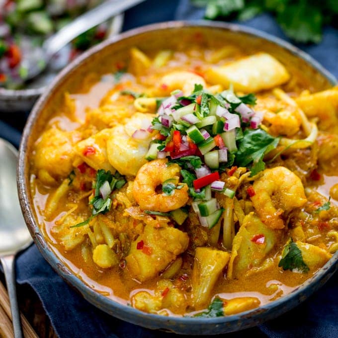 Square image of haddock and prawn laksa curry in a bowl with topping of chillies, cucumber and coriander. On a blue napkin, spoon on left hand side.