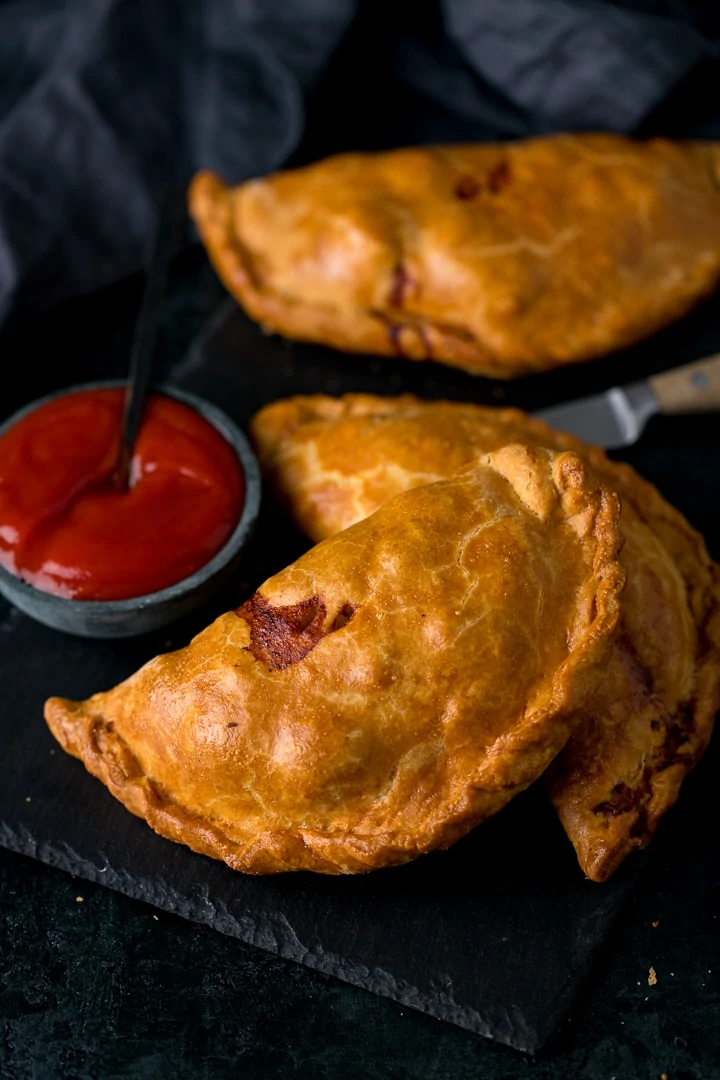 Cornish pasties on a dark serving board with ketchup next to them