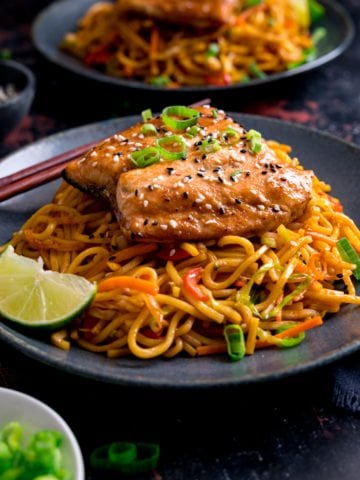 Square image of Asian salmon on top of stir fried noodles on a dark blue plate. Further plate of salmon and noodles in the background along with little plates of garnishes.