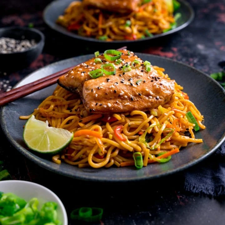 Square image of Asian salmon on top of stir fried noodles on a dark blue plate. Further plate of salmon and noodles in the background along with little plates of garnishes.