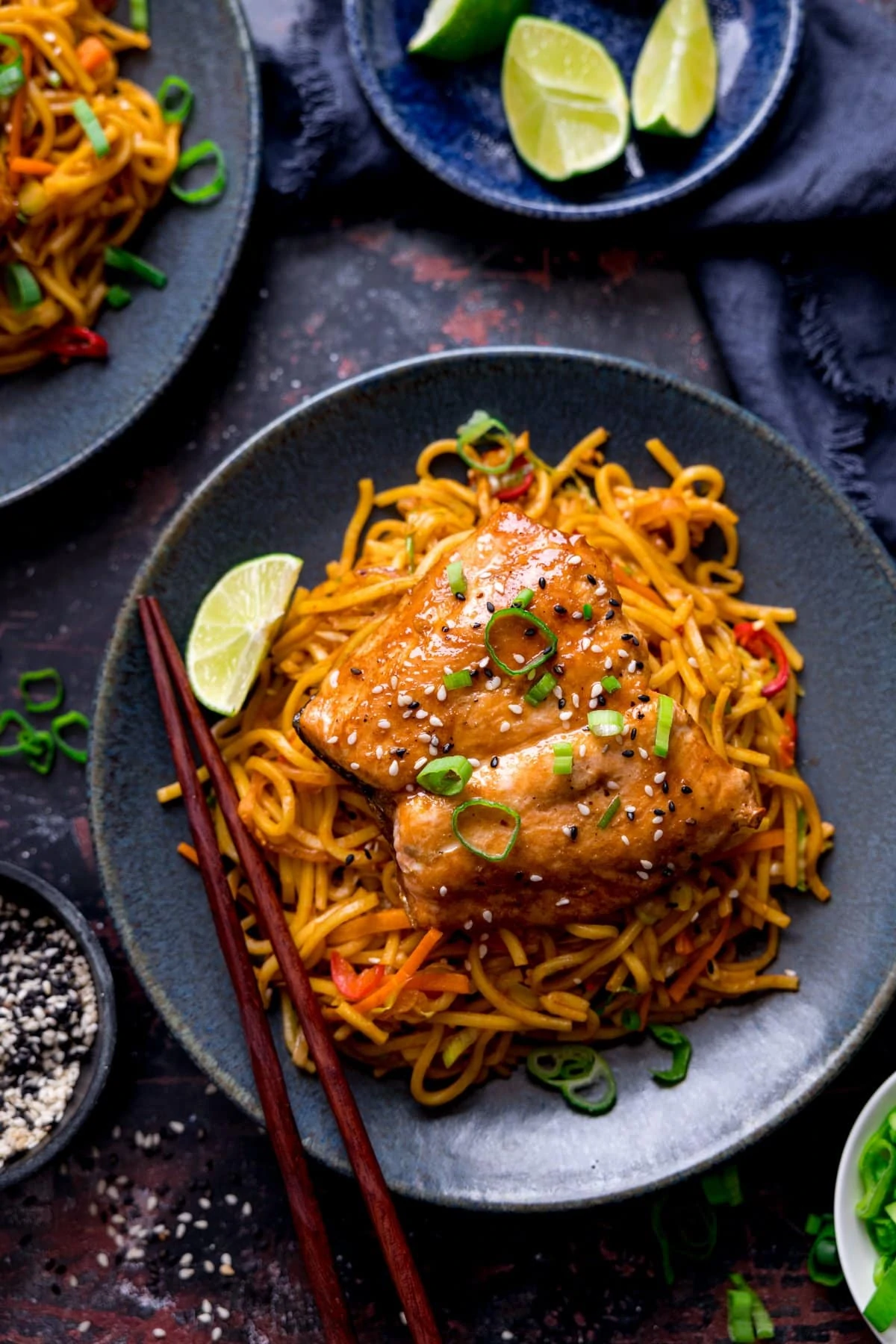 Overhead image of Asian salmon on a bed of noodles on a dark blue plate with lime wedge and chopsticks on the plate. The plate is on a dark background and there are bowls of garnishes around the central dish.