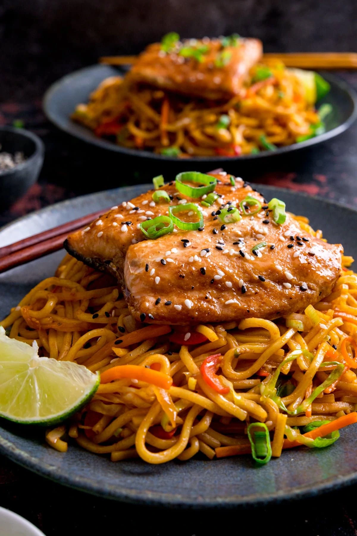 Asian salmon topped with spring onions on a bed of stir-fried noodles on a blue plate. Further plate with salmon and noodles in the background.
