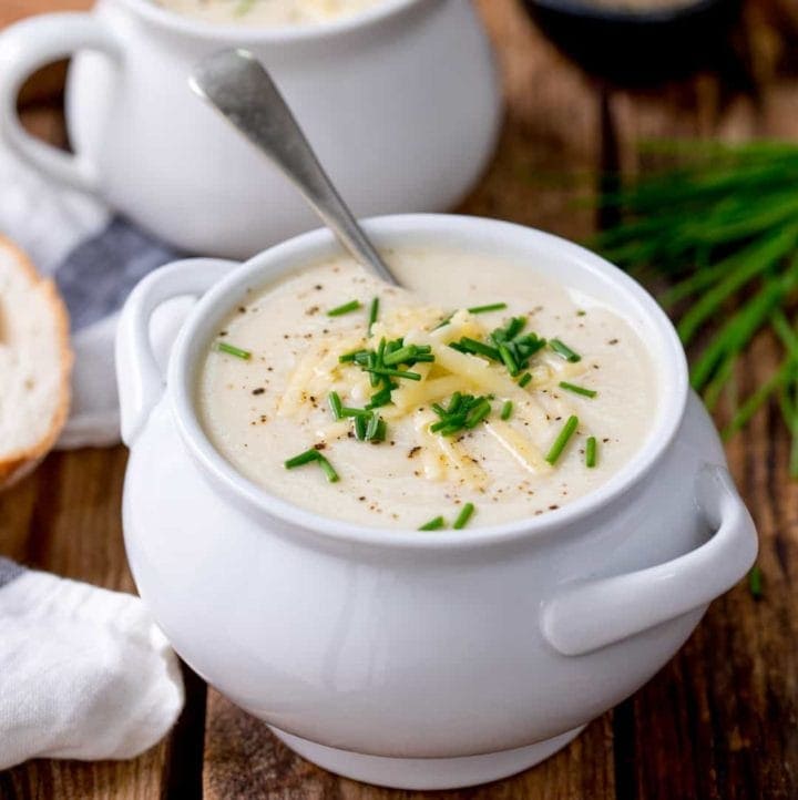 Cauliflower soup in a white bowl on a wooden background. Cheese and chives sprinkled on top of soup.