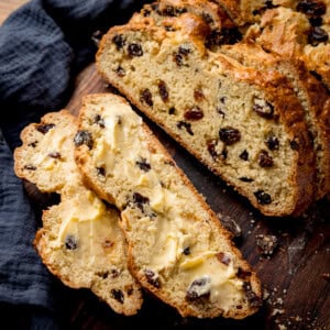 Sliced fruit soda bread with butter rests on a wooden surface, accompanied by a dark cloth.