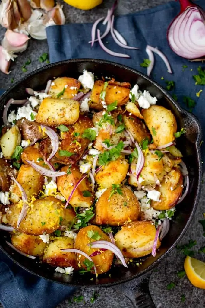 Overhead shot of crispy Greek style potatoes in a pan with feta, red onions, parsley and lemon zest. Ingredient scattered around.