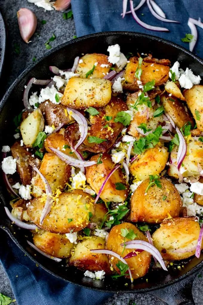 Pan of fried Greek potatoes with feta, red onion and parsley on a blue background.