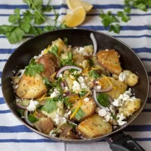 Pan of Greek potatoes with feta, red onion and parsley on a blue and white striped tea towel