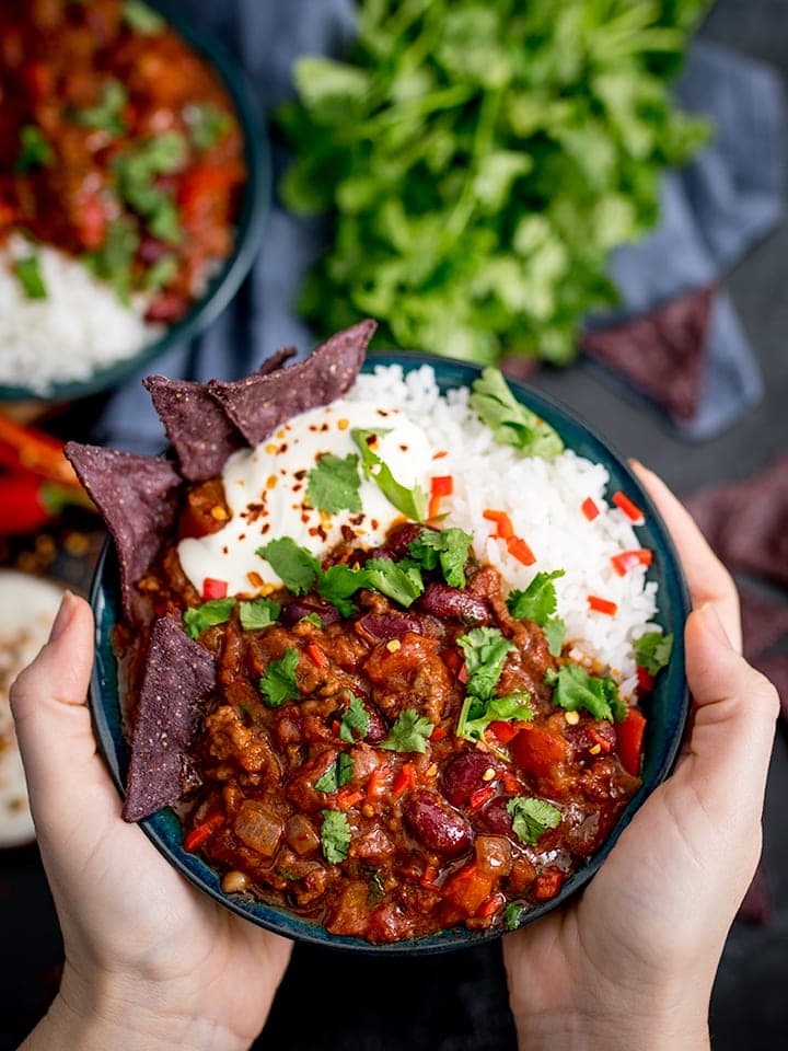 Hands holding bowl of Bowl of chilli con carne with rice and blue corn chips