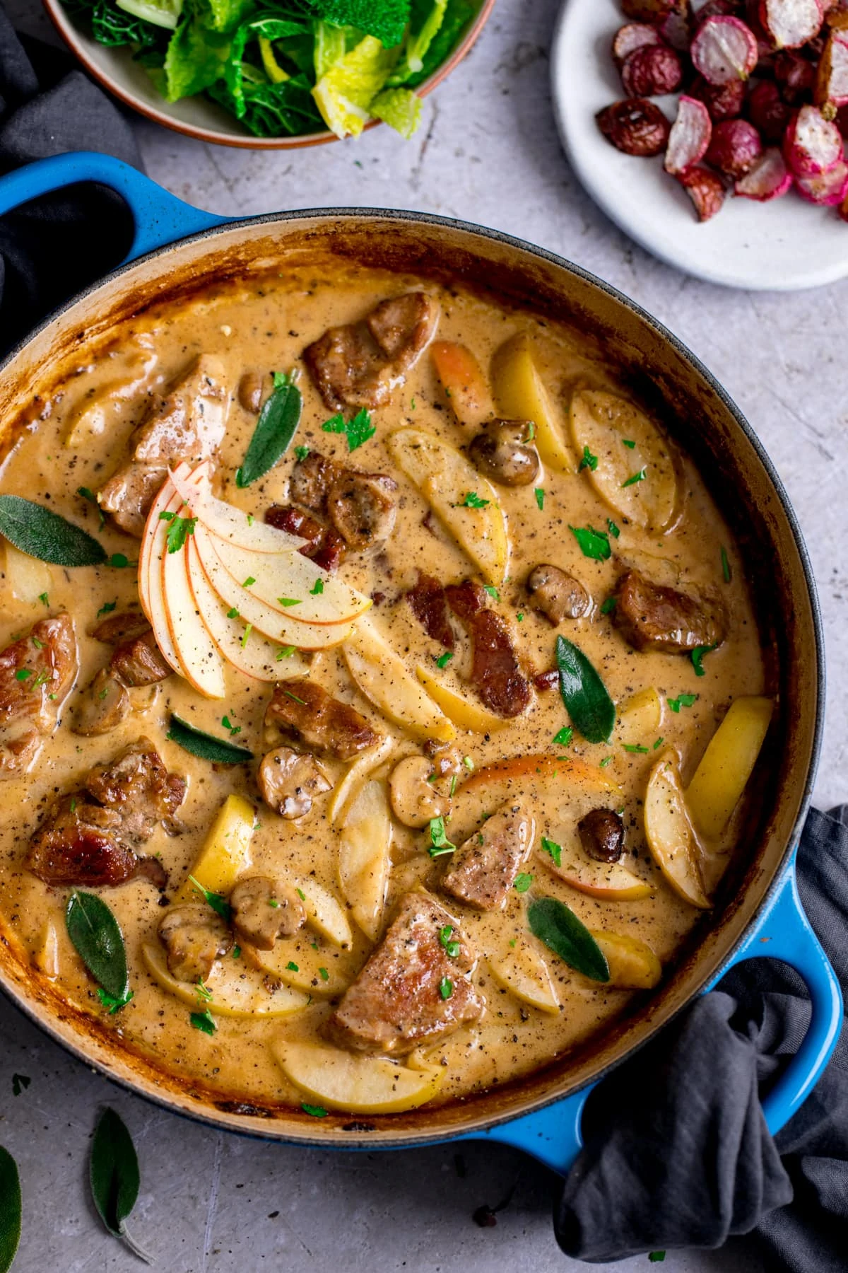 Overhead image of creamy pork and apple casserole in a blue casserole dish on a light background. Side dishes of cabbage and radishes are just in shot.