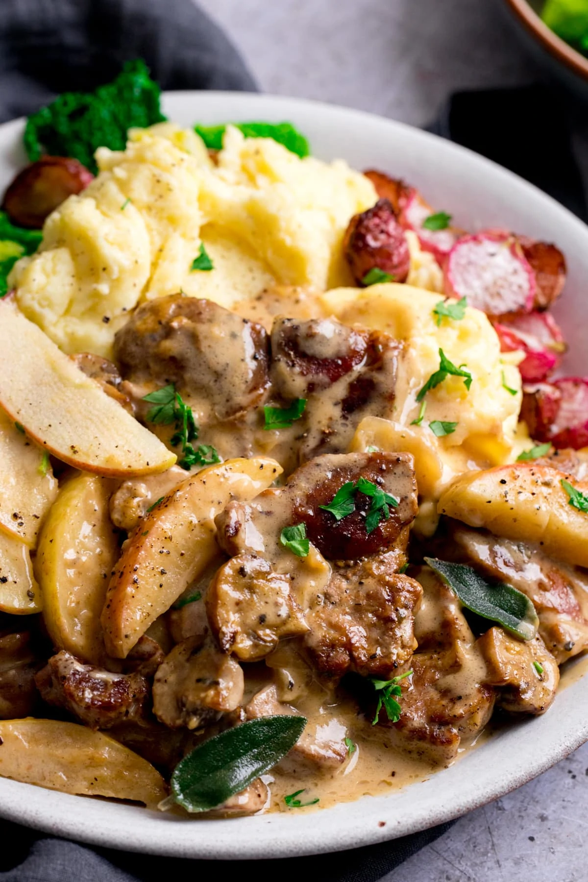 Close up of creamy pork and apple casserole on a white plate with mashed potatoes and roasted radishes.