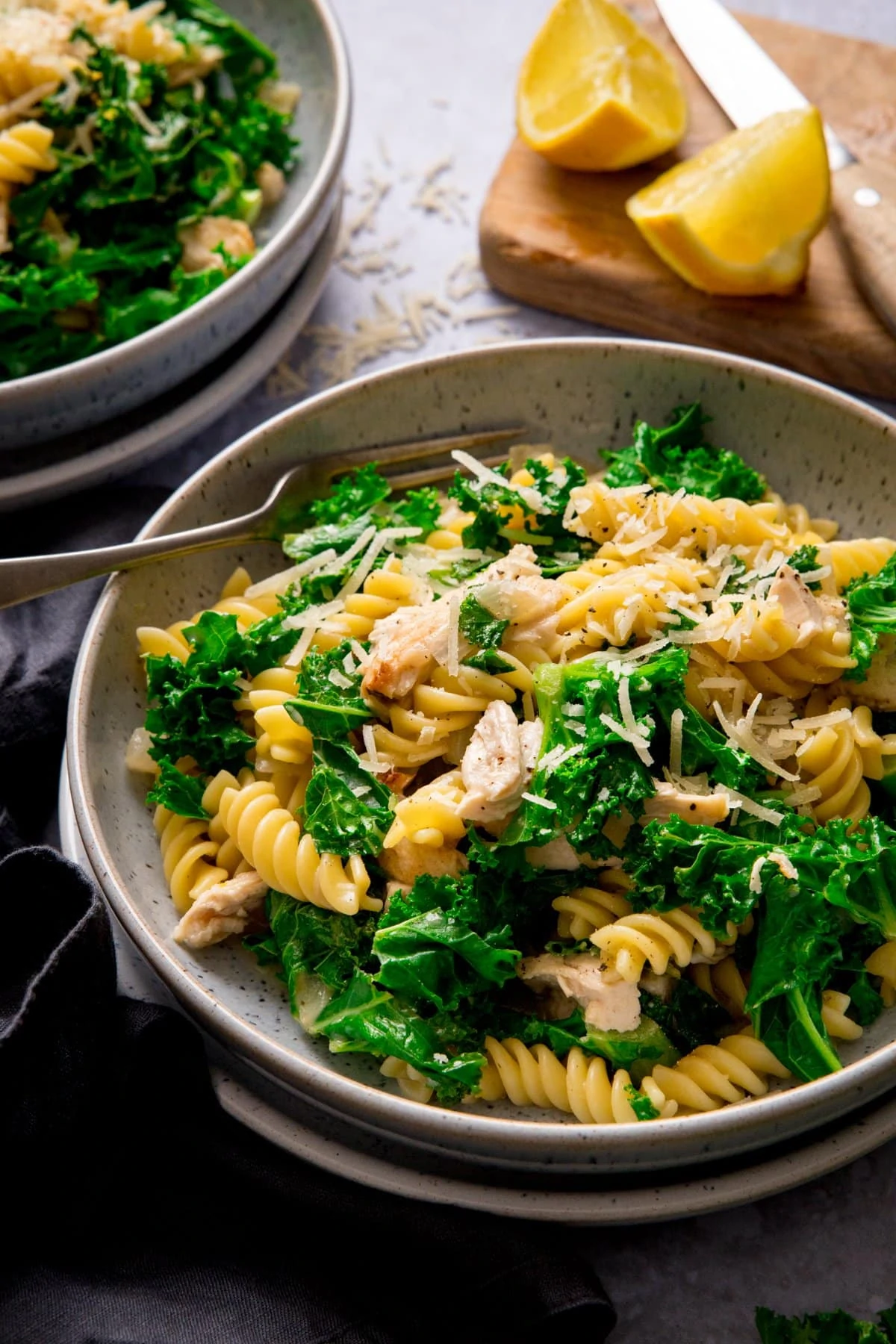 Bowl of garlic chicken pasta and kale with a fork in. Wedges of lime and further bowl of pasta at the top of the shot