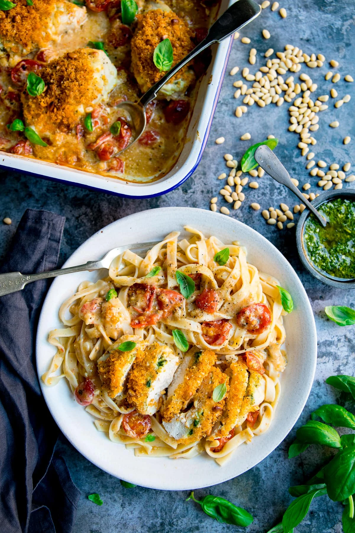 baked pesto chicken on a white plate with tagliatelle pasta. Dish of pesto chicken just in shot. Dishes are on a blue background.