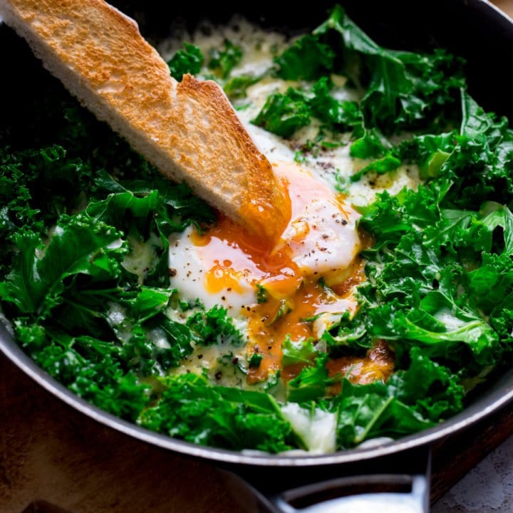 A toast soldier being dipped into a runny egg yolk in a pan of egg and cheesy kale
