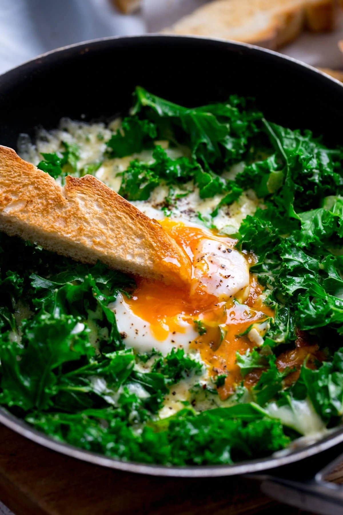 A toast soldier being dipped into the runny egg yolk in a pan of kale and fried egg.
