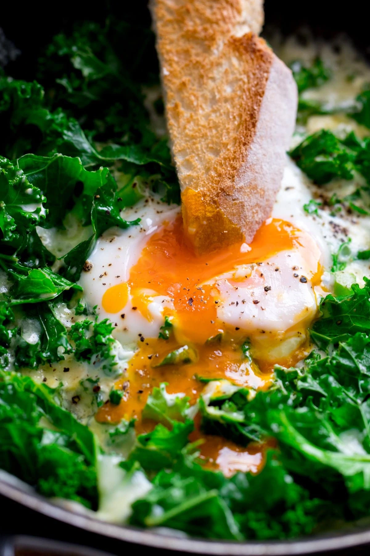 Close up of a toast soldier being dipped into a runny egg in a pan of egg and cheesy kale