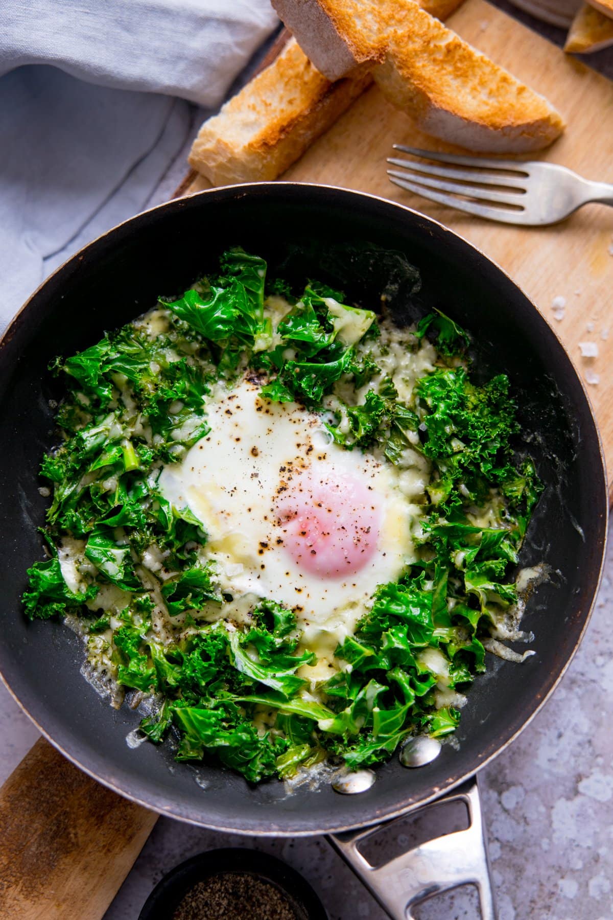 Fried egg in a pan with kale and cheese. Pan is on a wooden board with toast soldiers at the top of the shot.