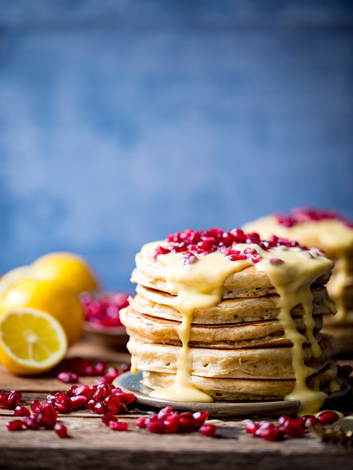 Stack of ricotta pancakes topped with lemon sauce and pomegranate with a blue backdrop