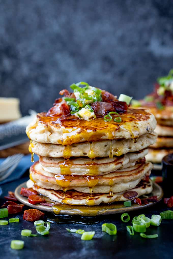 Stack of savoury pancakes against a dark background. Pancakes are topped with bacon, chilli butter and spring onions.