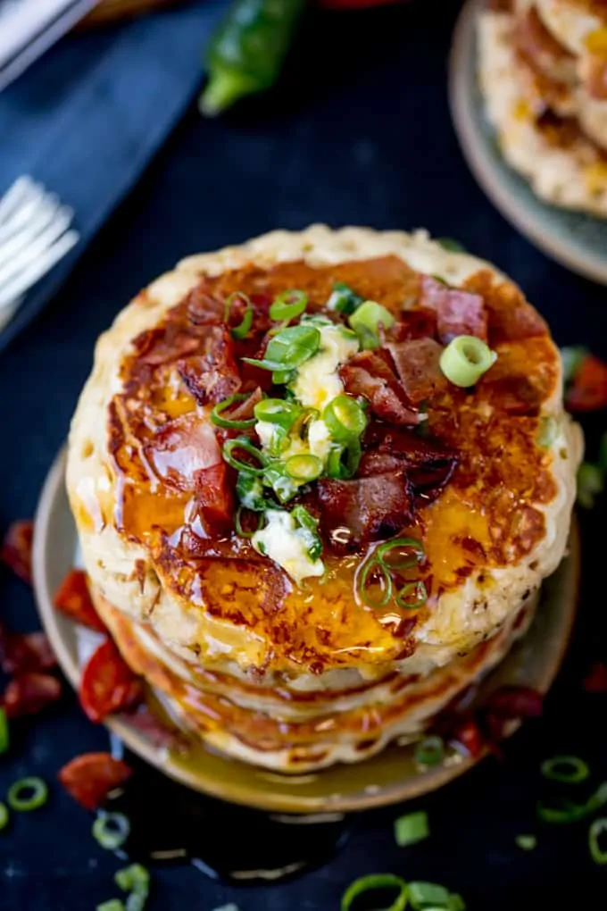 Overhead image of a stack of savoury pancakes with bacon, spring onions and chilli butter on a dark background