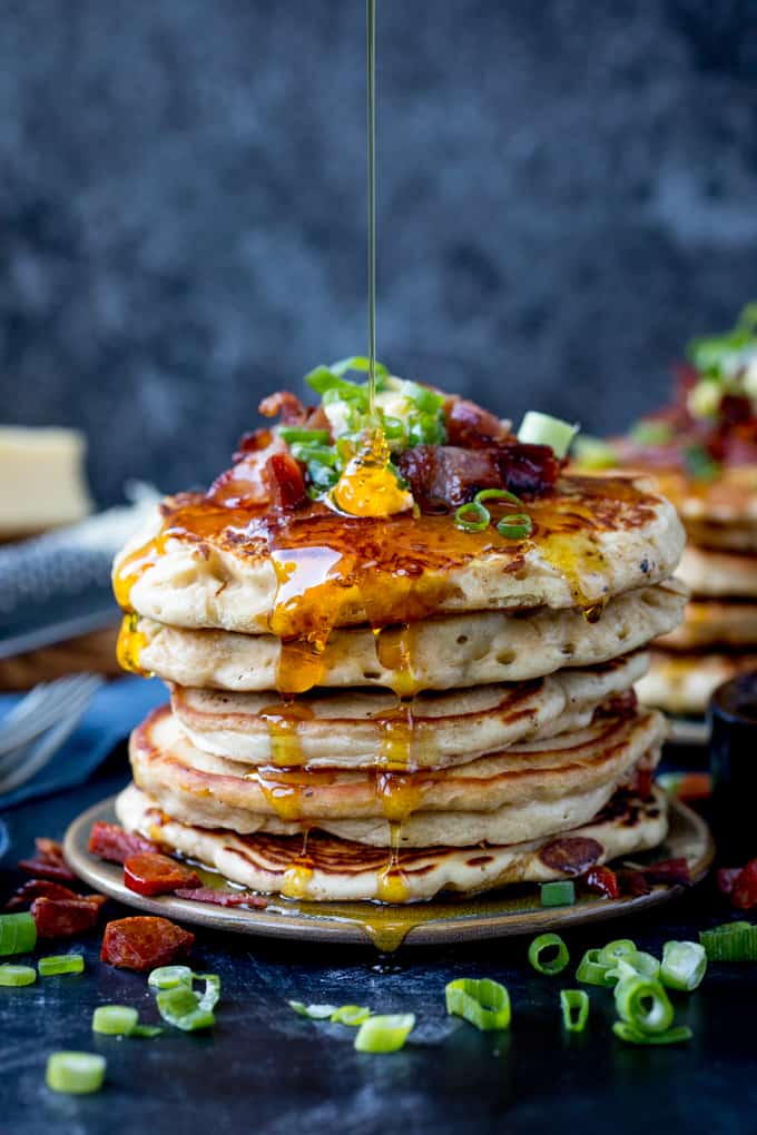 Savoury pancakes stacked up on a plate and topped with bacon, chilli butter and spring onions. Golden syrup being drizzled on.