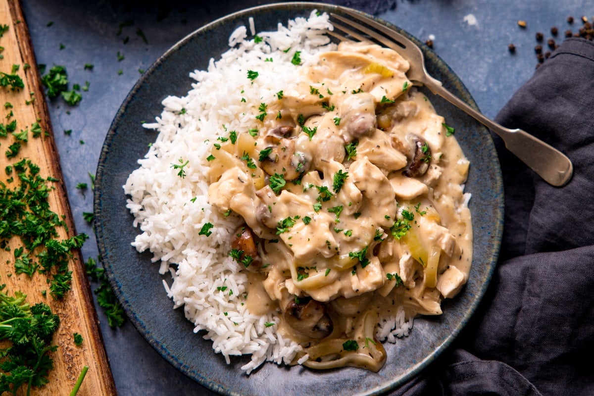 Wide image of chicken ala king with boiled rice on a dark plate on a blue backgrouns. Chopping board with parsley on just in shot to the left.