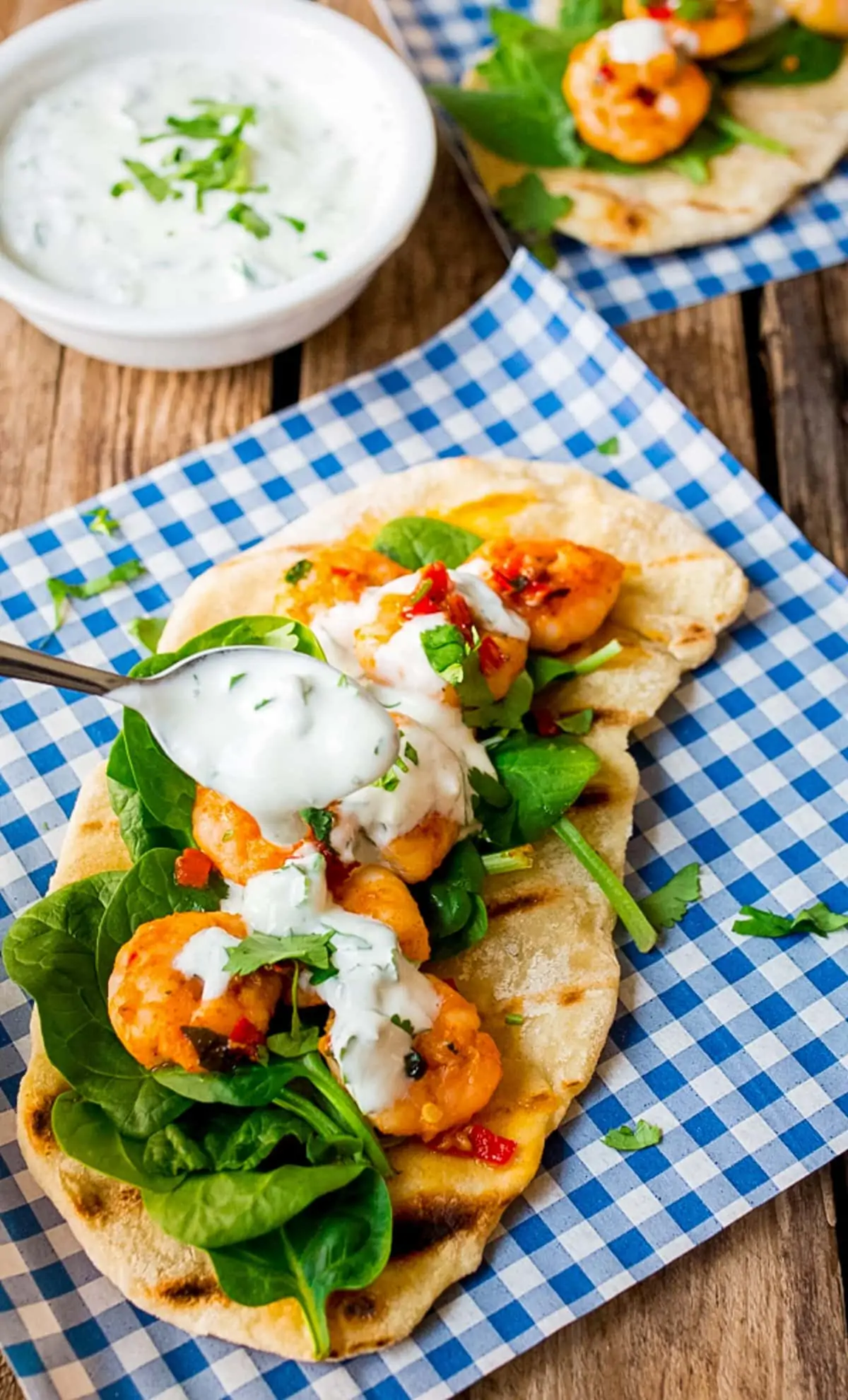 Spoonful of yogurt dip being drizzled on garlic chilli prawns with spinach on homemade flatbread on a piece of blue chequered paper