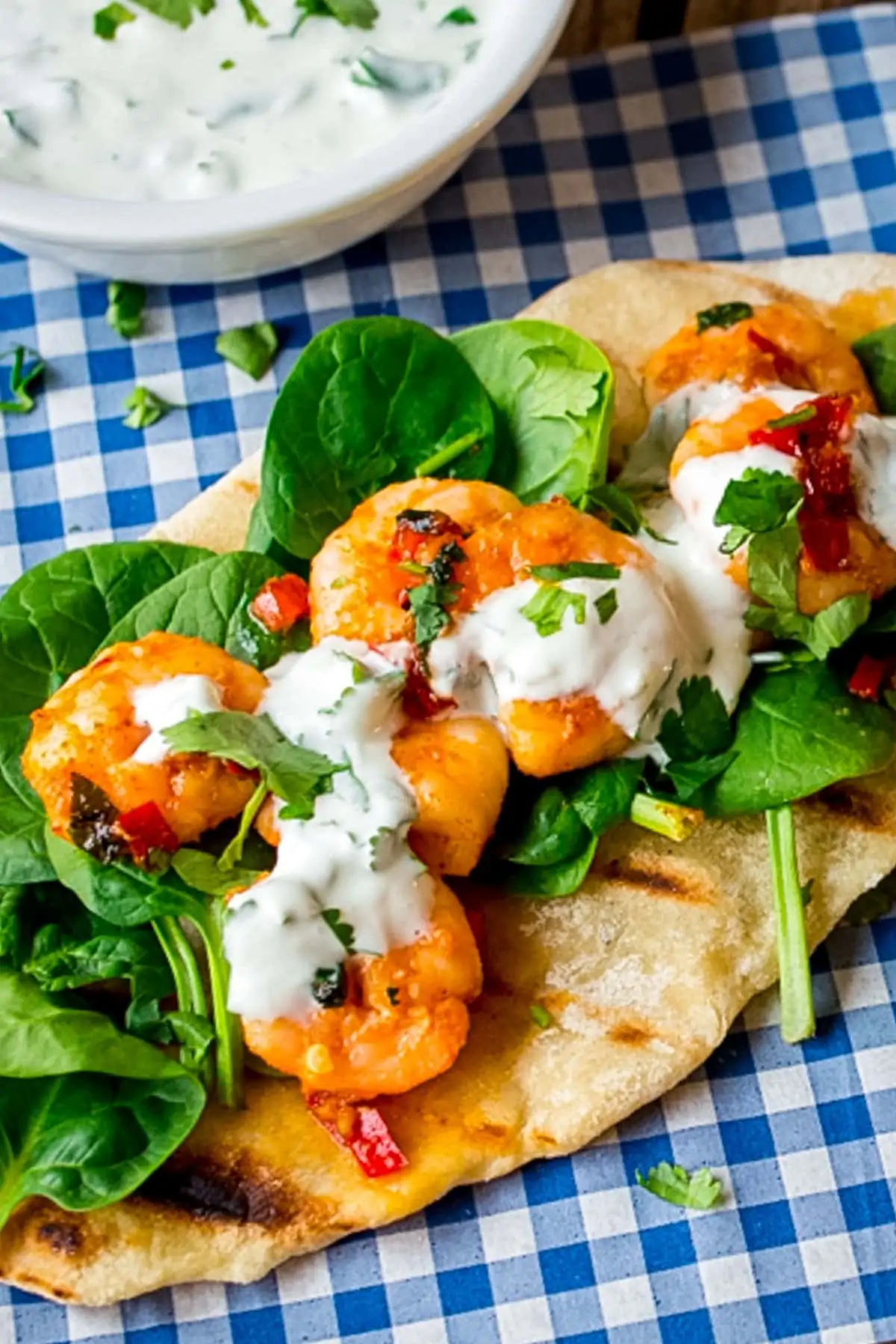 Tall image of Garlic chilli prawns with spinach and yogurt dip on homemade flatbread on a piece of blue chequered paper