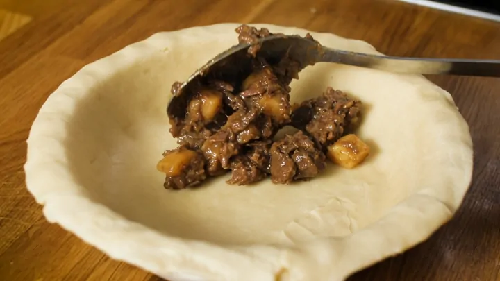 Steak and potato filling being spooned into pie base