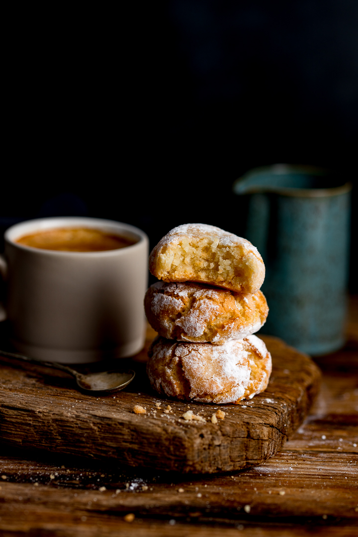 Tall image of amaretti cookies on a wooden board against a dark background. Coffee and milk jug in background.