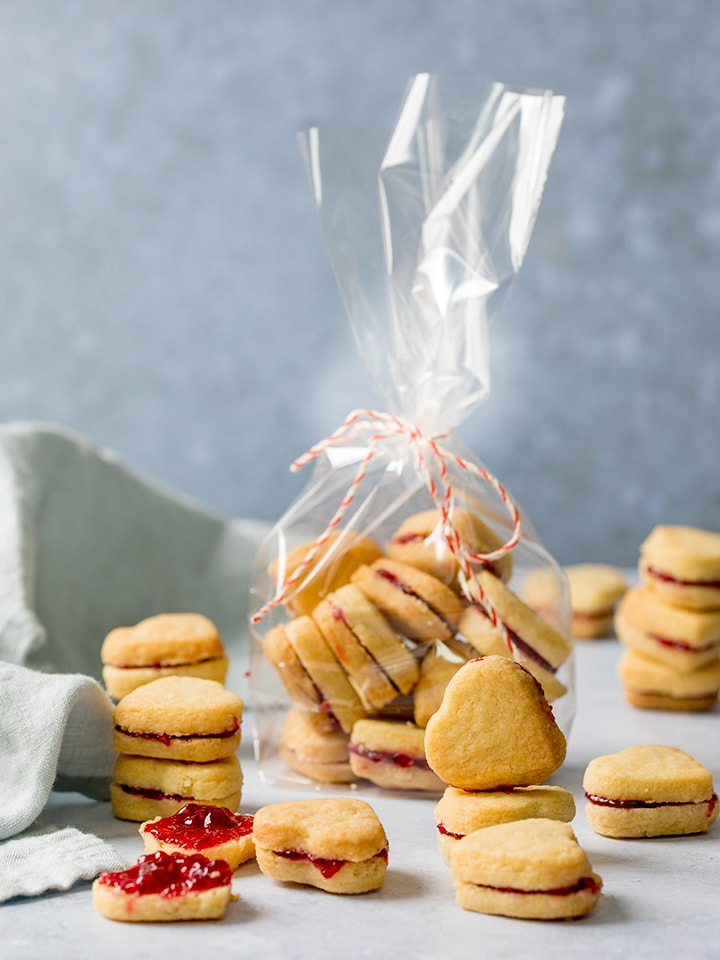 Stack of jam-filled, heart-shaped valentine shortbread cookies, plus some in a gift bag on a light blue background