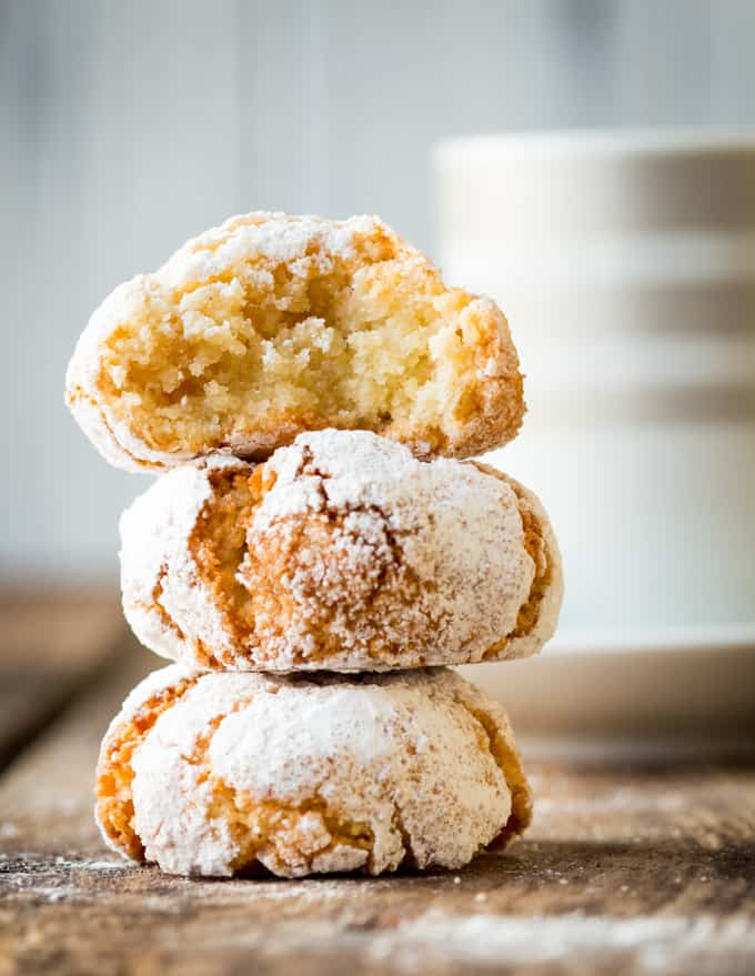 3 Italian Amaretti cookies in a pile on a wooden board. The top one has a bite taken out. Jar of cookies and a coffee in the background.