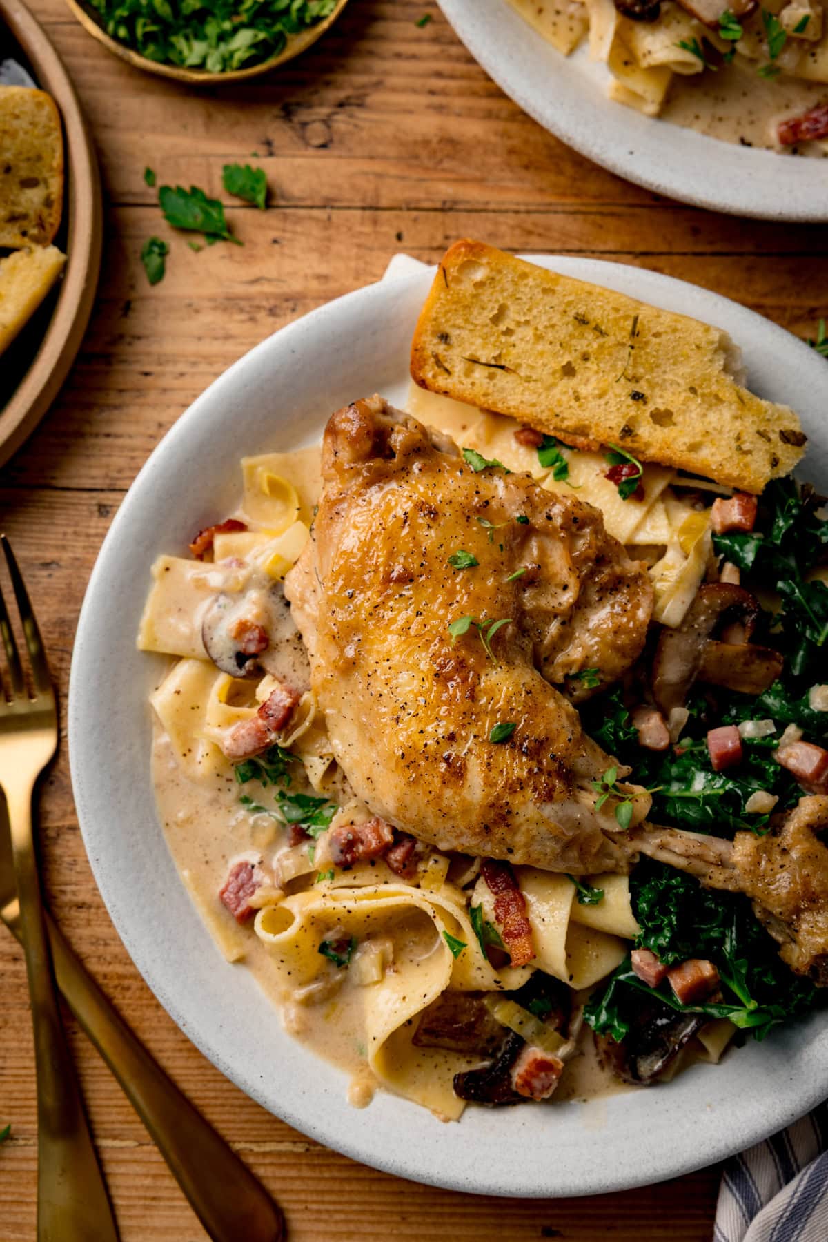 A white plate of coq au vin blanc with pappardelle pasta, kale and garlic bread on a wooden surface. There is a further plate, cutlery and chopped parsley in shot.